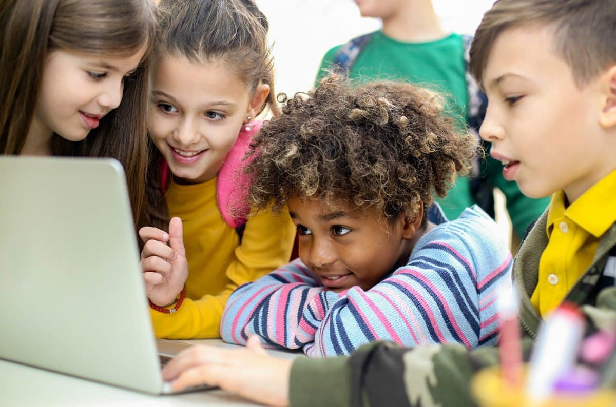 Group of diverse young children smile together and look at a laptop screen on a table.