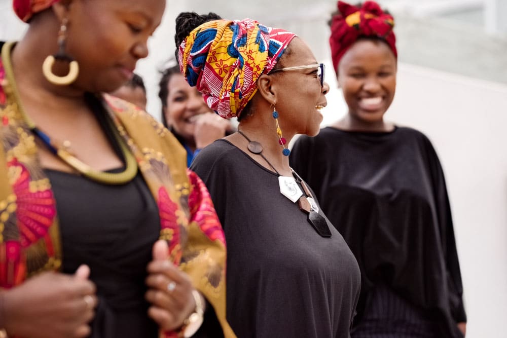 A small group of African women wearing black business attire and colourful headwraps smile and talk to each other