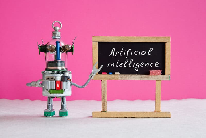 Tiny robot made from metal spare parts points to a tiny chalkboard with the words "Artificial Intelligence" written on it